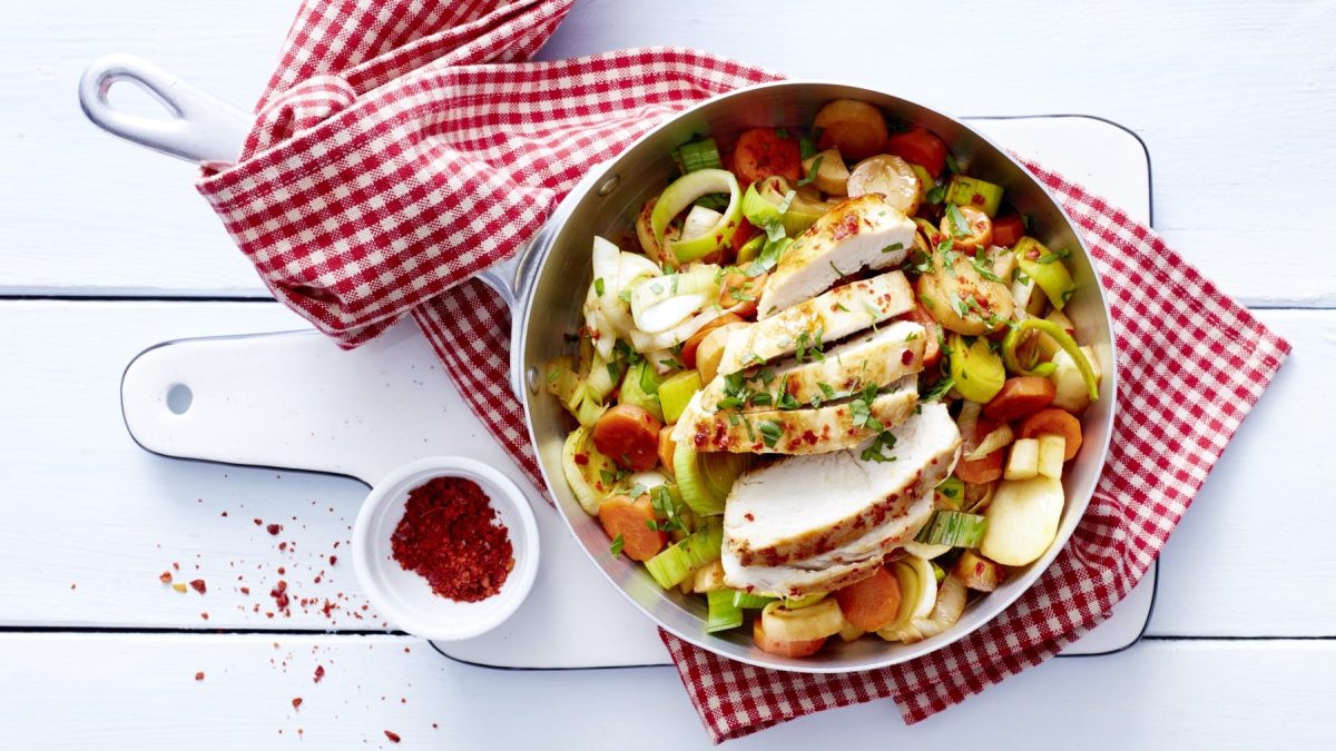 Blitz-Gemüse mit Huhn in einer Pfanne mit einem rot-weiß-karierten Küchentuch auf einem Schneidenbrett, daneben ein weißes Schälchen mit Chiliflocken. Alles in der Draufsicht vor weißem Hintergrund.