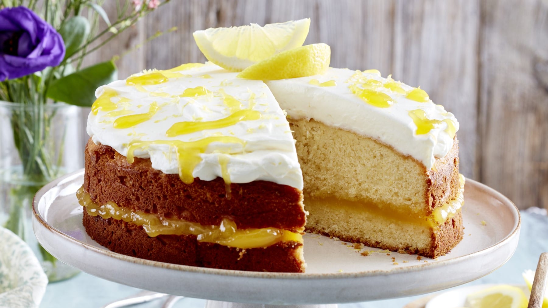 Angeschnittener Lemon-Curd-Kuchen mit Sahne und Zitronenspalten auf einem Servierteller, Holzwand und blaue Blumen im Hintergrund.