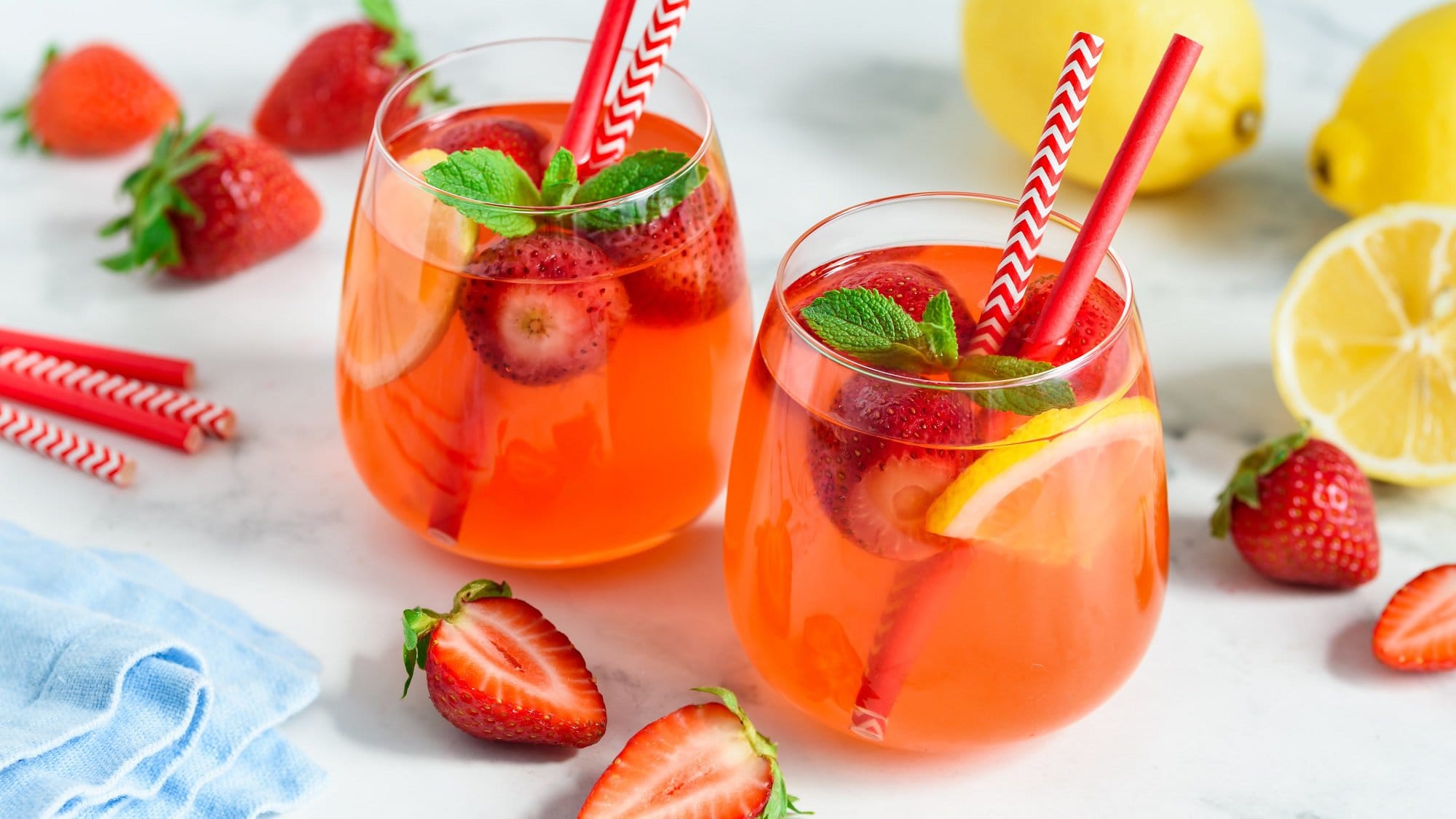 Schräge Draufsicht: Zwei Gläser mir einem erfrischenden Drink mit Erdbeeren und Zitronen stehen auf einem hellen Untergrund. Dahinter liegen drei Zitronen sowie einige Erdbeeren.
