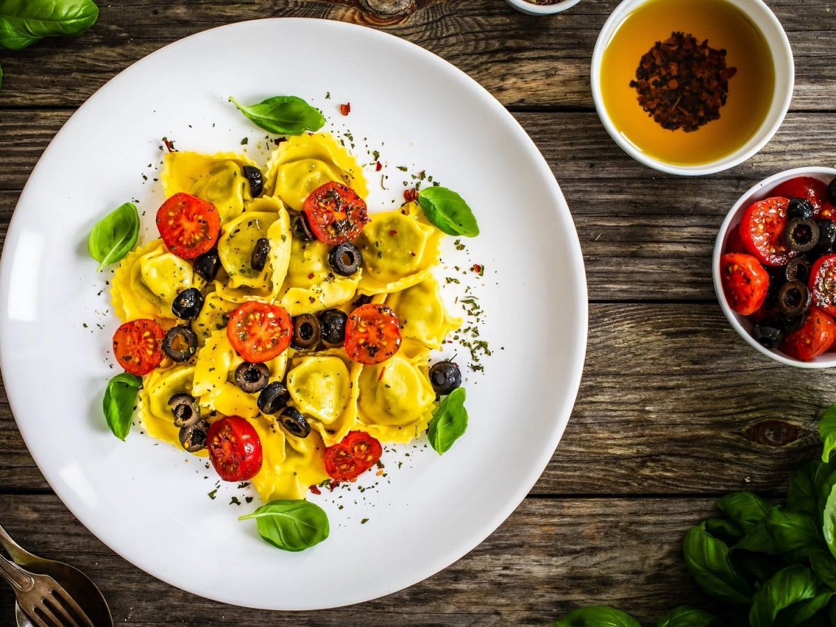 Draufsicht: Ein Teller Gefüllte Tortellini mit Oliven und Tomaten auf einem Holztisch. Daneben liegt frisches Basilikum, sowie zwei Schälchen mit Gewürzölen.