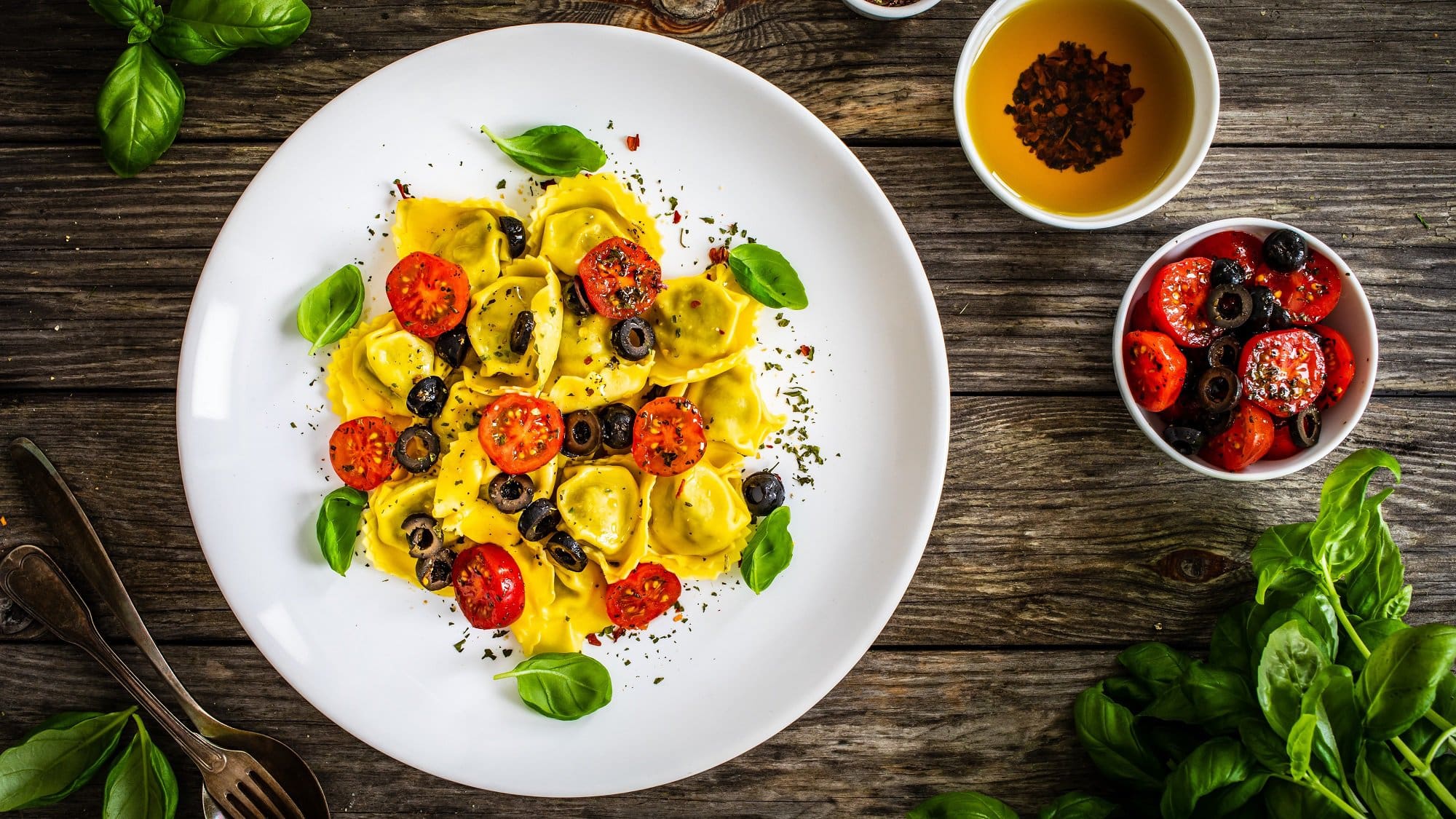 Draufsicht: Ein Teller Gefüllte Tortellini mit Oliven und Tomaten auf einem Holztisch. Daneben liegt frisches Basilikum, sowie zwei Schälchen mit Gewürzölen.