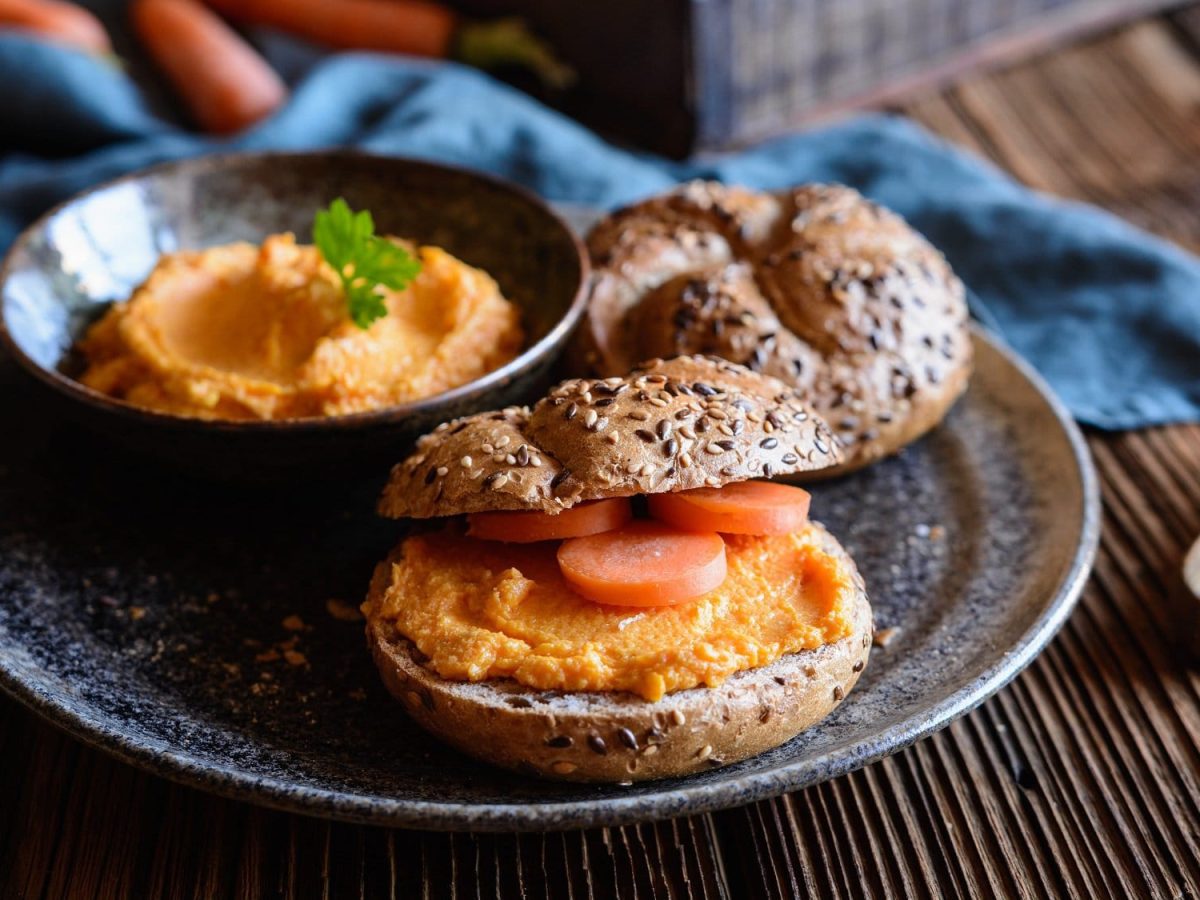 Ein Brötchen mit Karotten-Hummus und Möhrenscheiben auf einem Teller, darauf auch eine Schale vom Hummus.