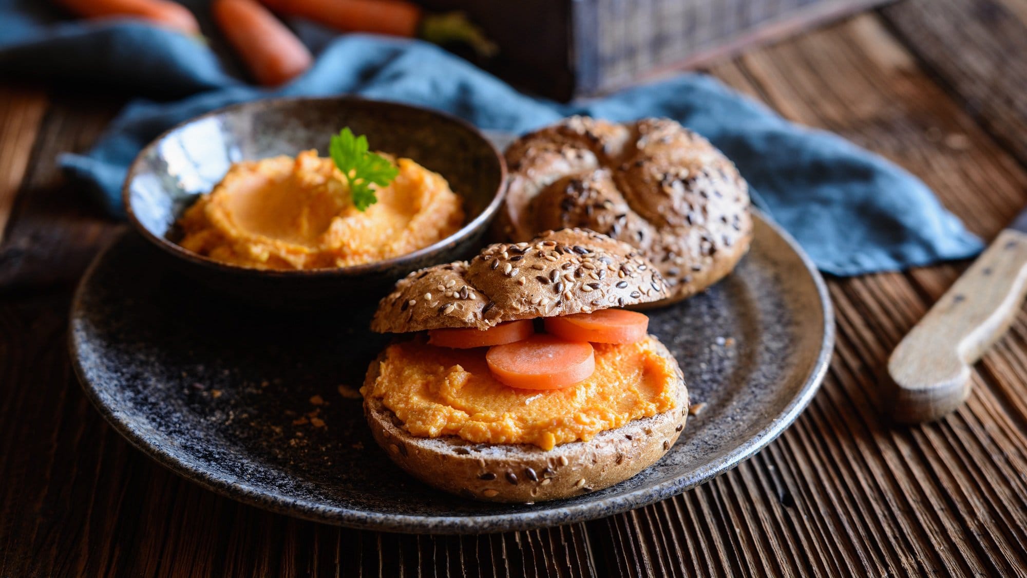 Ein Brötchen mit Karotten-Hummus und Möhrenscheiben auf einem Teller, darauf auch eine Schale vom Hummus.