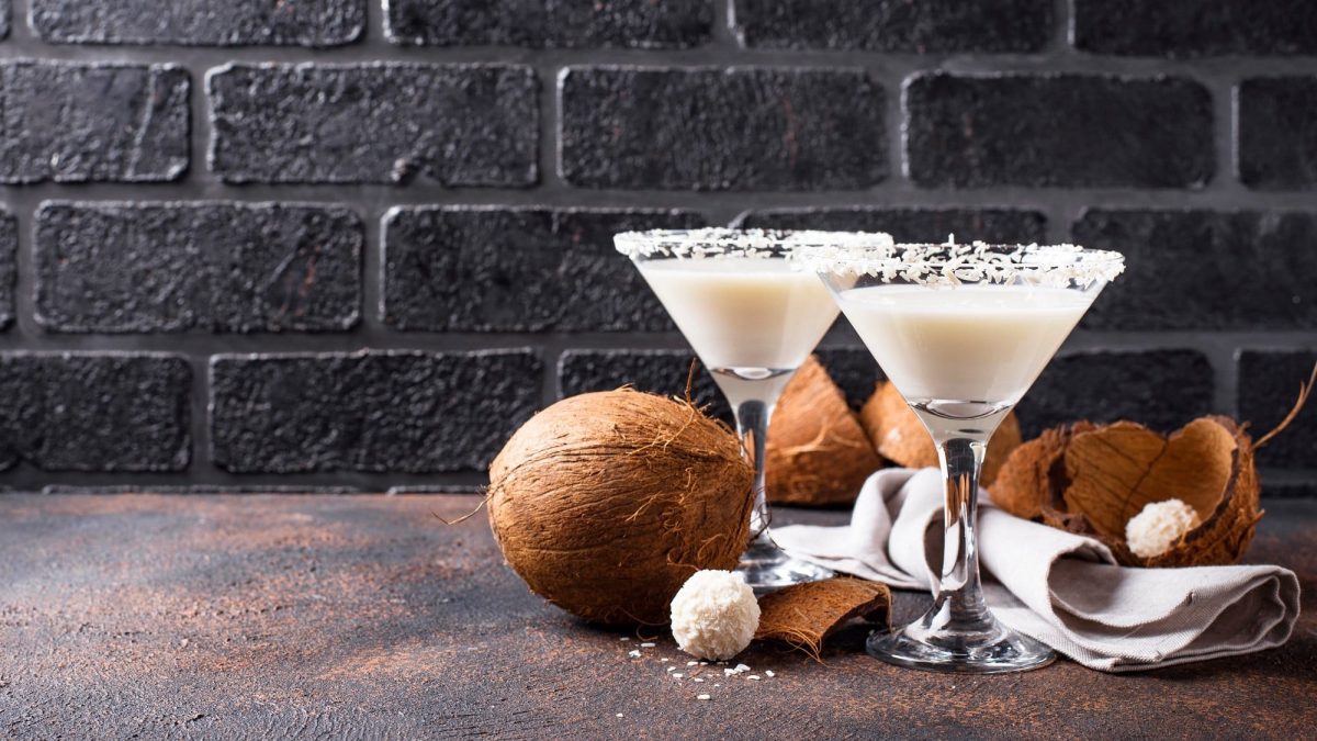Zwei Gläser Kokos-Margarita mit Kokosraspeln verziert, drumherum liegen Kokosnüsse und Kokospralinen.