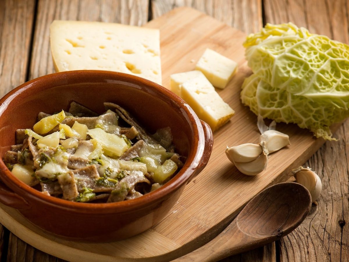 Eine braune Schale mit den Pizzoccheri-Nudeln auf einem Holzbrett mit Käse, Knoblauchzehen und etwas Wirsing.