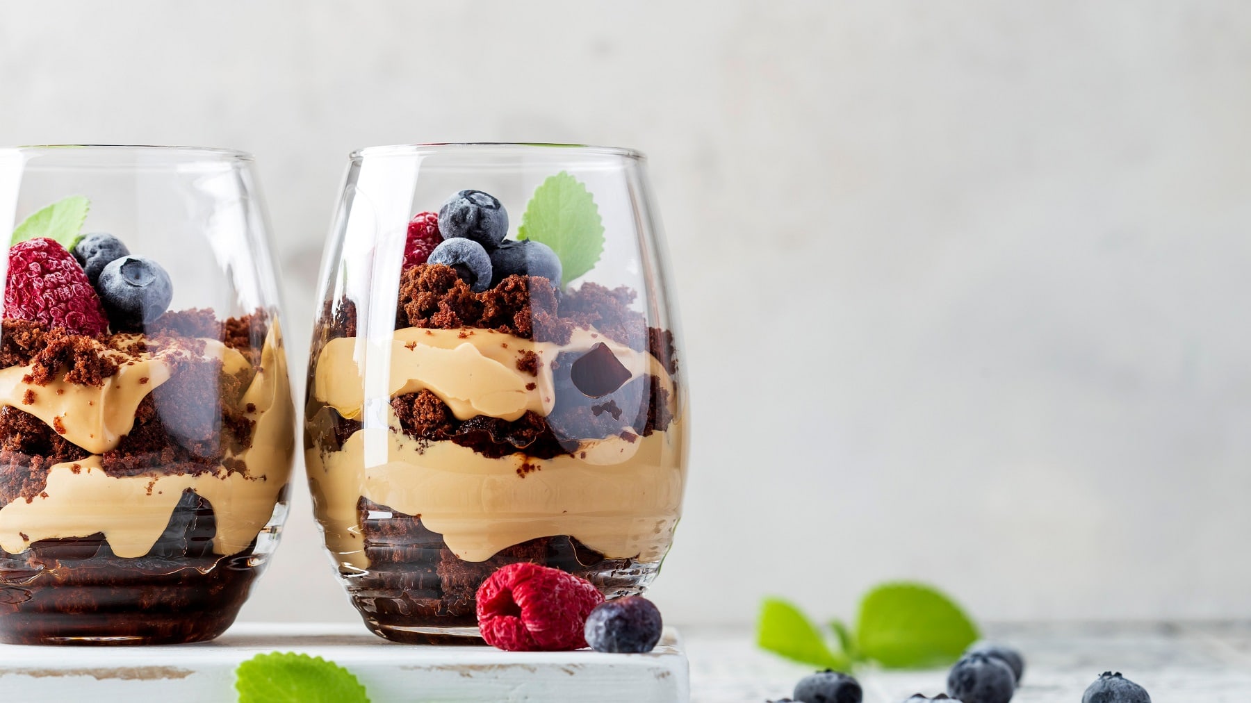 Zwei Gläser mit dem Schoko-Beeren-Dessert auf einem hellen Tisch mit Heidelbeeren und Zitronenmelisse.