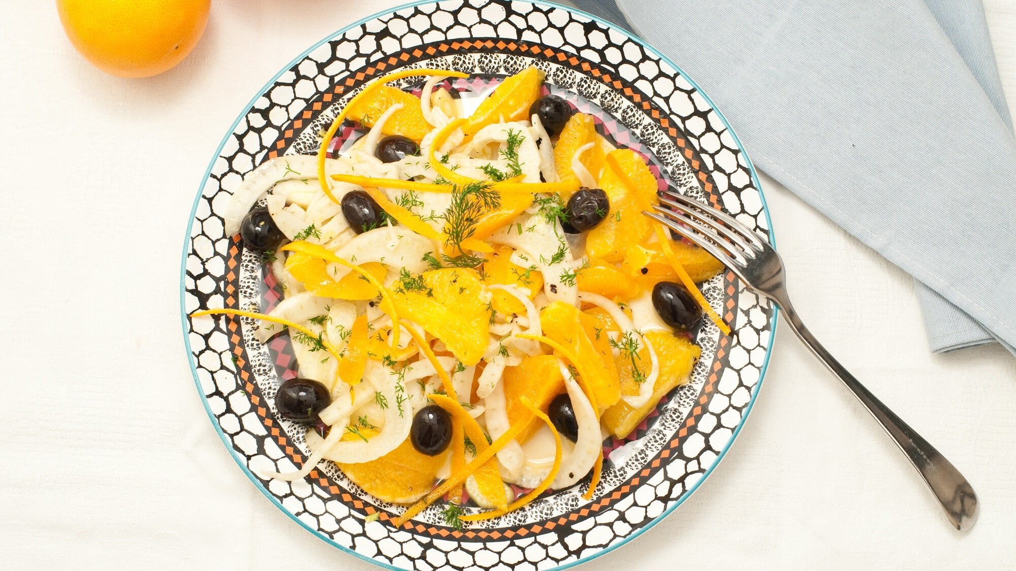 Draufsicht: ein mediterran bemalter Teller mit einem sizilianischem Fenchel-Oliven-Salat mit Mandel-Dressing steht auf einem weißen Untergrund. Daneben liegen eine Gabel und ein hellblaues Tuch.