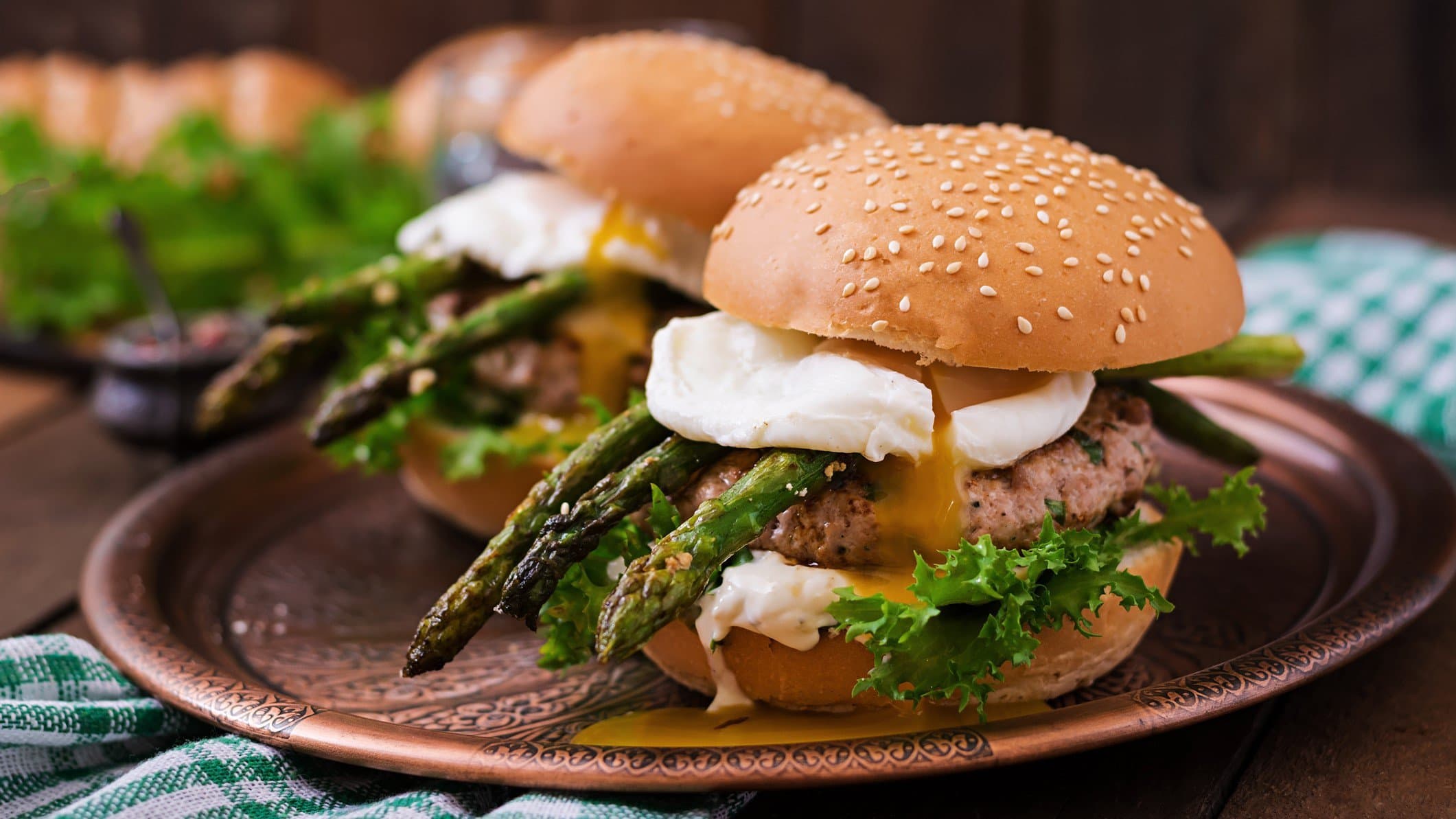 Zwei Spargel-Burger mit pochiertem Ei, Chicken-Patty, grünem Spargel und Burrata auf braunem Teller. Im Hintergrund weitere Zutaten. Frontalansicht.