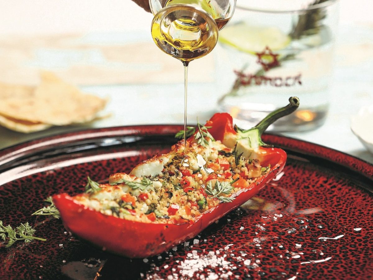 Eine halbe Spitzpaprika mit Gemüse-Couscous-Füllung auf einem Teller, darüber wird etwas Öl gegossen.