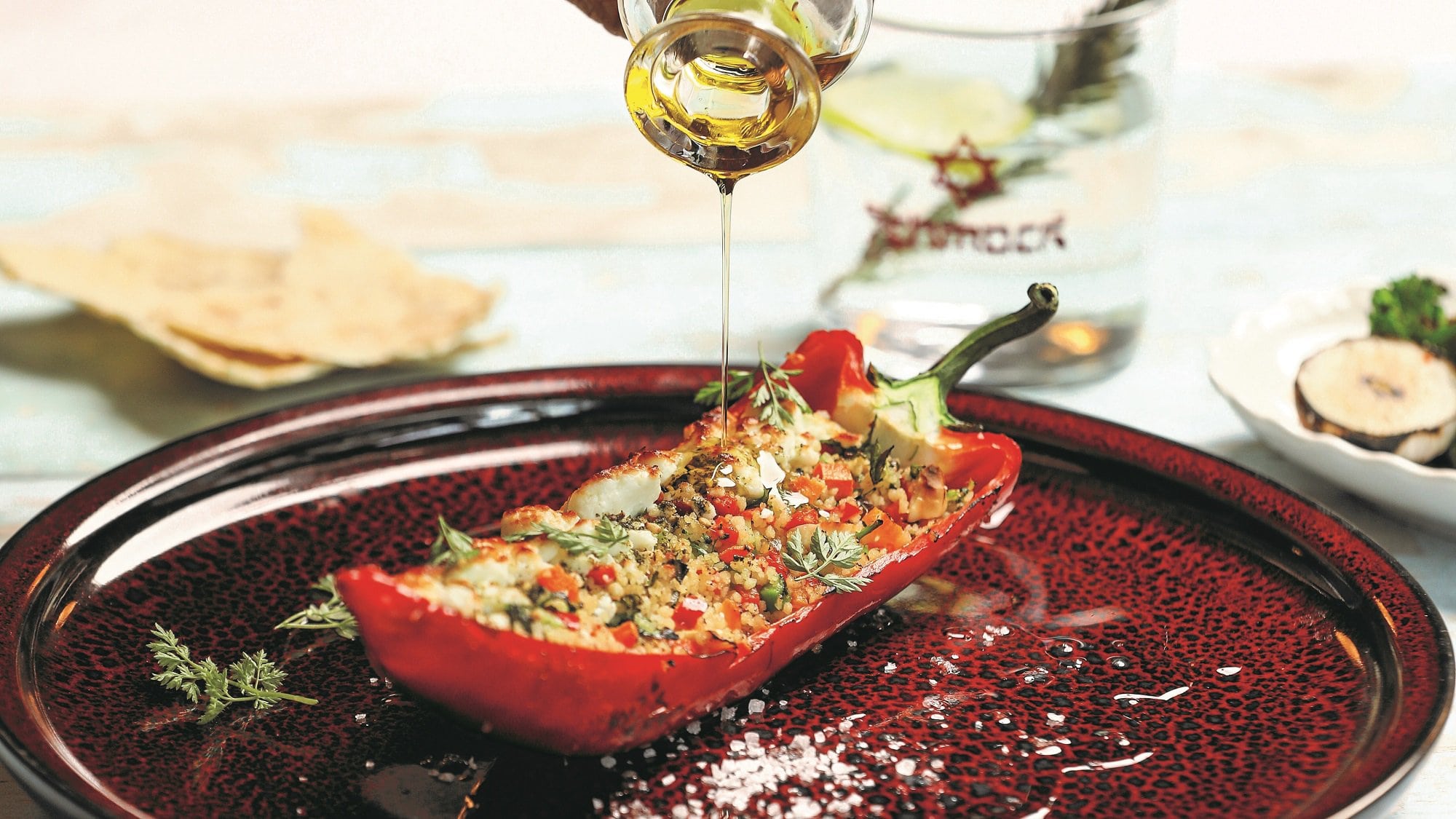 Eine halbe Spitzpaprika mit Gemüse-Couscous-Füllung auf einem Teller, darüber wird etwas Öl gegossen.