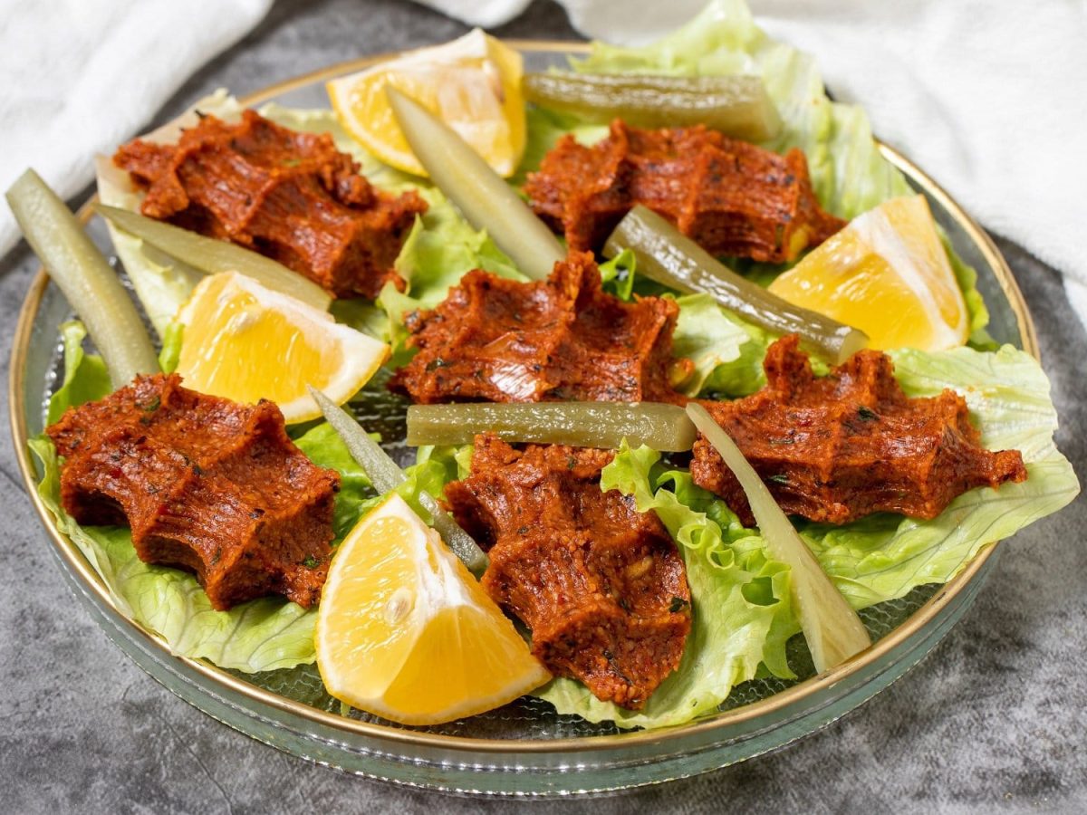 Vegane Çiğ Köfte auf Salatblättern mit Zitronenstücken und in Streifen geschnittener Gewürzgurken auf einem silbernen Teller.