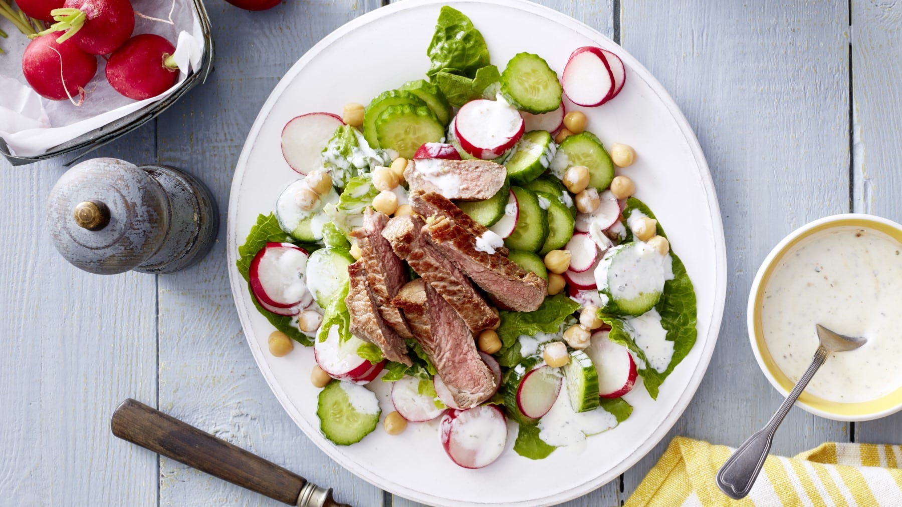 Draufsicht: Zaziki-Salat mit Steakstreifen auf einem weißen Teller. Außerdem eine Schüssel mit weißer Sauce, Gewürzmühle und Radieschen vor hellem Holzhintergrund.