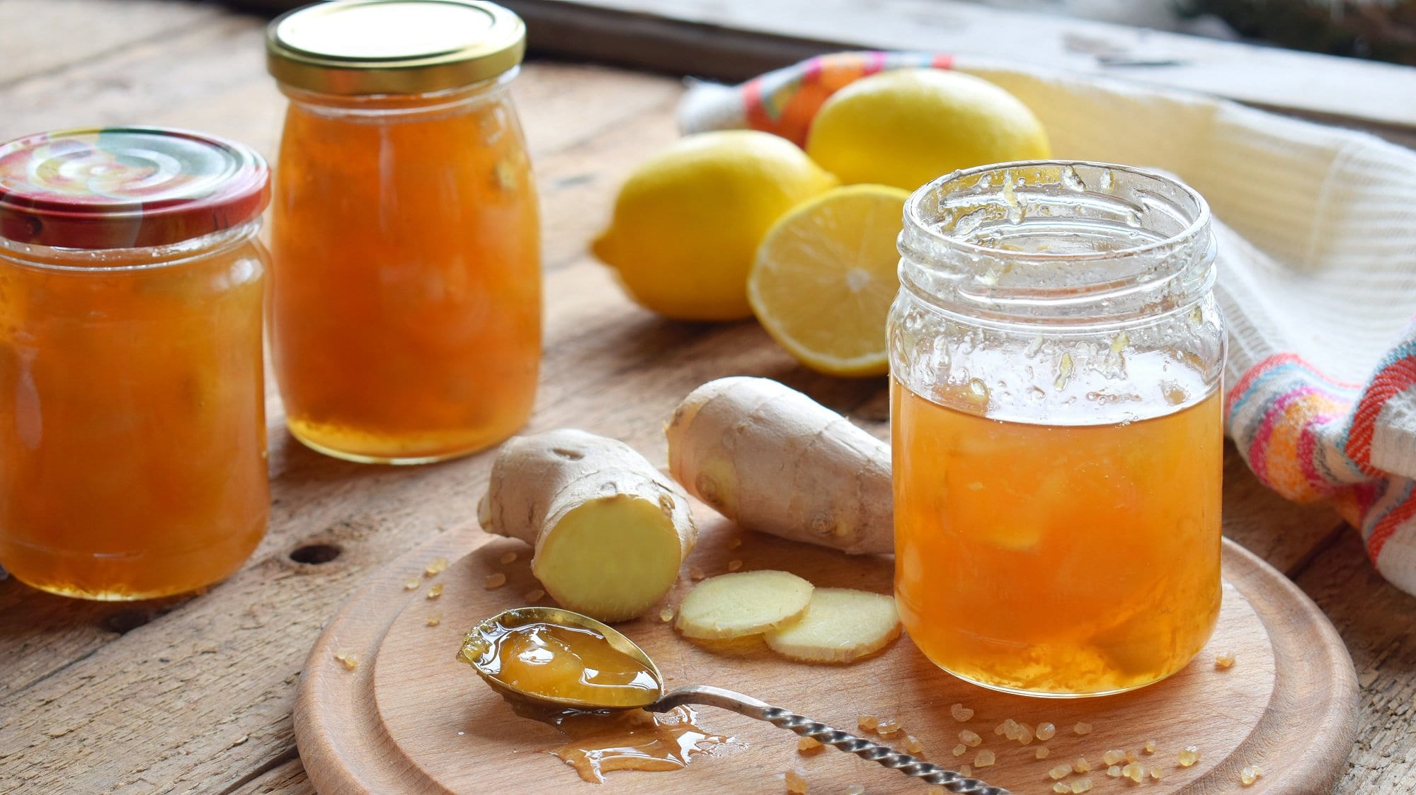 Frontal: drei Gläser Zitronen-Ingwer-Marmelade stehen auf einem Untergrund aus Holz. Drum herum liegen frischer Ingwer und Zitronen und ein Teelöffel.
