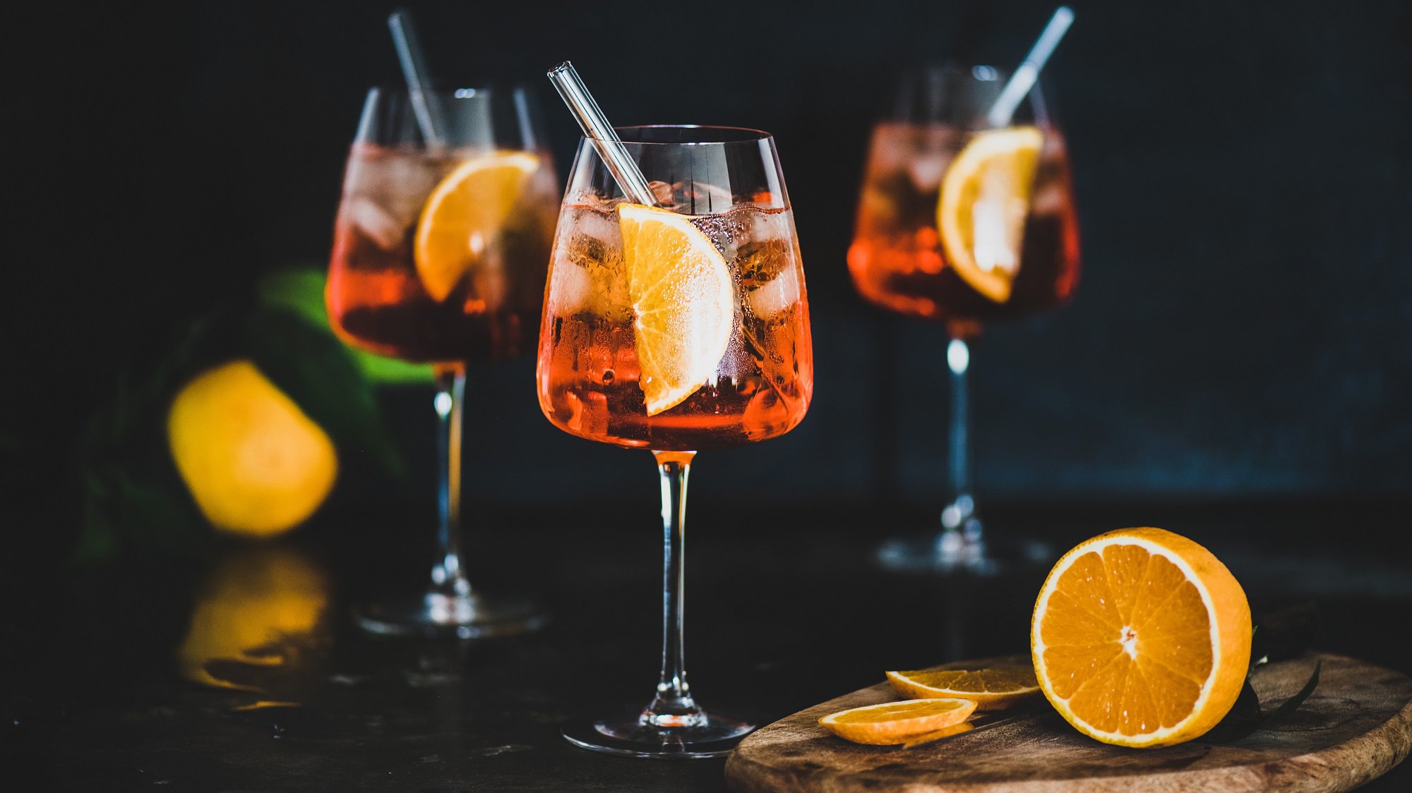 Drei Gläser alokoholfreier Aperol Spritz mit je einem Strohhalm und einer Orangenscheibe darin, daneben eine angeschnittene Orange.