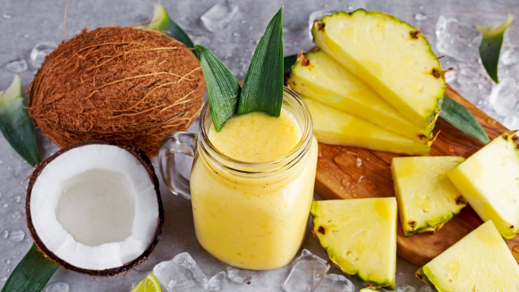 Ein Glas mit dem Ananas-Kokos-Smoothie auf einem grauen Tisch mit den beiden Früchten und Eiswürfeln.