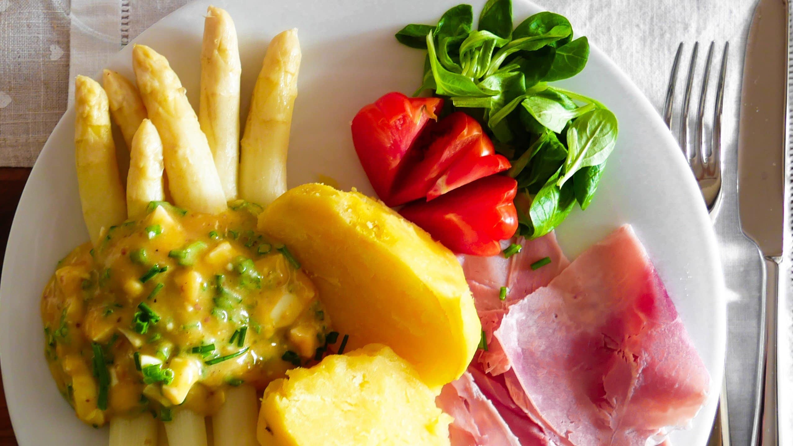 Asparagi alla Bolzanina Spargel mit Bozner Sauce auf weißem Teller mit Kartoffeln, Schinken und Salat. Draufsicht.