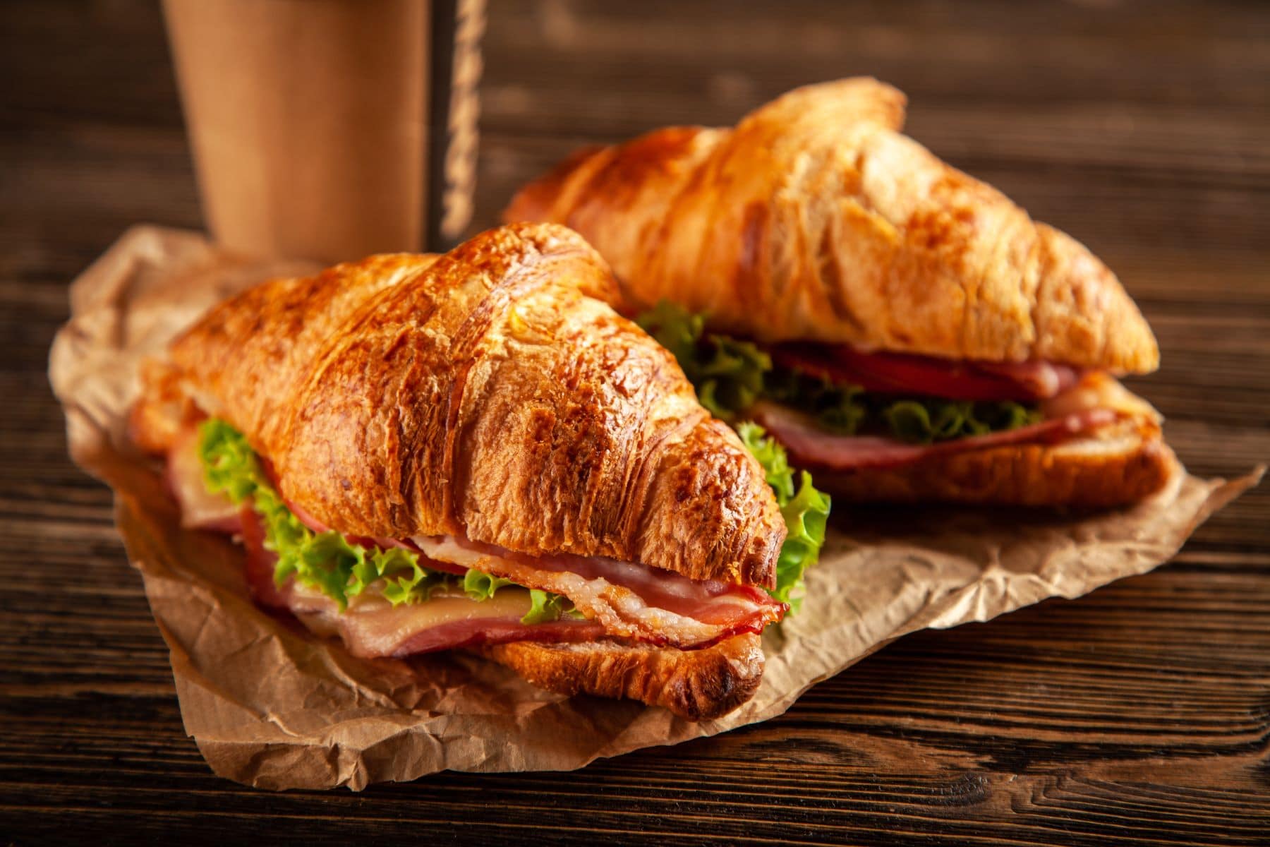 Zwei BLT-Croissant-Sandwiches auf Papier mit einem To-Go-Kaffeebecher auf Holz. Draufsicht.