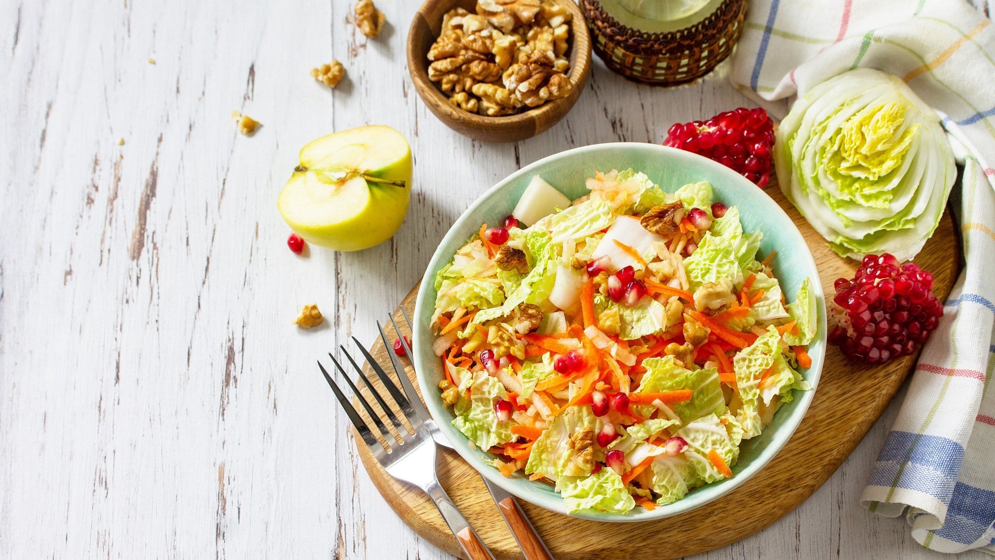Draufsicht: Auf einem weißen Untergrund aus Holz steht eine Schüssel Chinakohl-Salat mit Granatapfelkernen. Drumherum liegen einige der Zutaten.