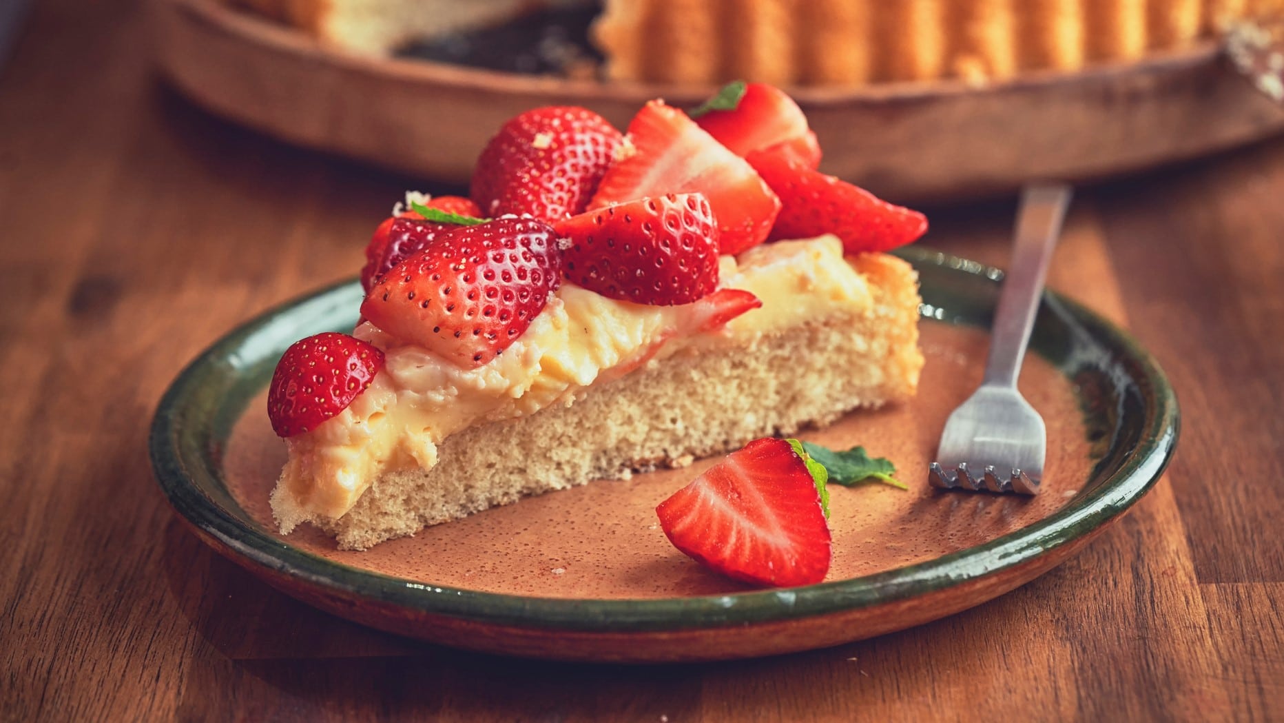 Ein Stück Erdbeer-Pudding-Kuchen mit Kuchengabel auf Teller auf Holz. Im Hintergrund angedeutet Tarte-Form. Frontalansicht.