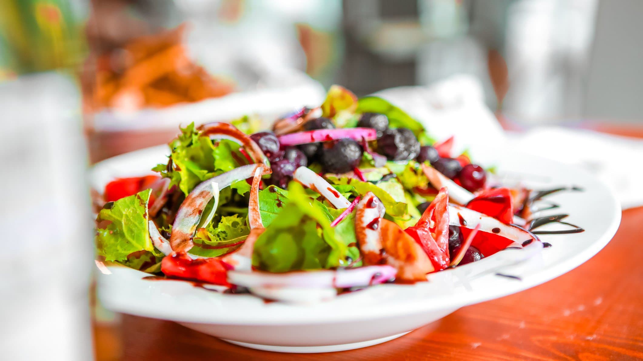 Frischer Frühlingssalat mit Tomaten, roten Zwiebeln, Oliven und Erdbeer-Vinaigrette in weißem Teller auf rotem Untergrund. Frontalansicht