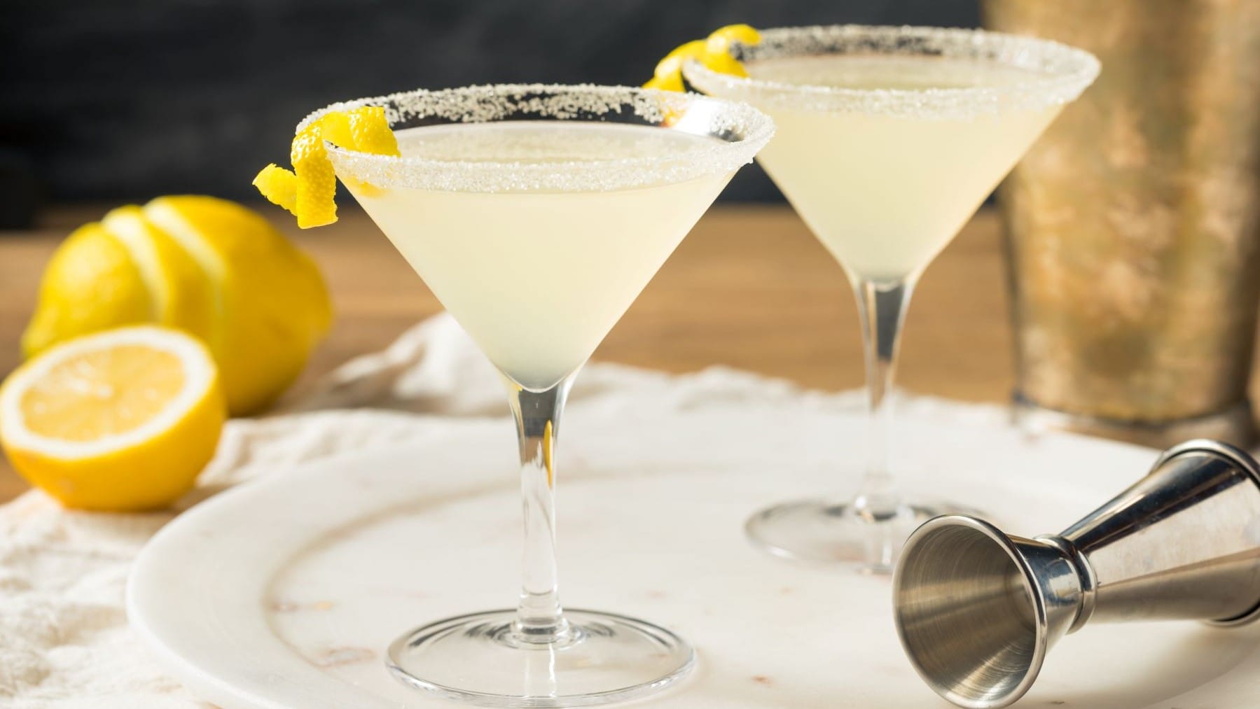 Zwei Lemon Drop Martini mit Barmaß auf hellem Teller, Tuch und Holztisch. Im Hintergrund Shaker und Zitronen. Frontalaufnahme.