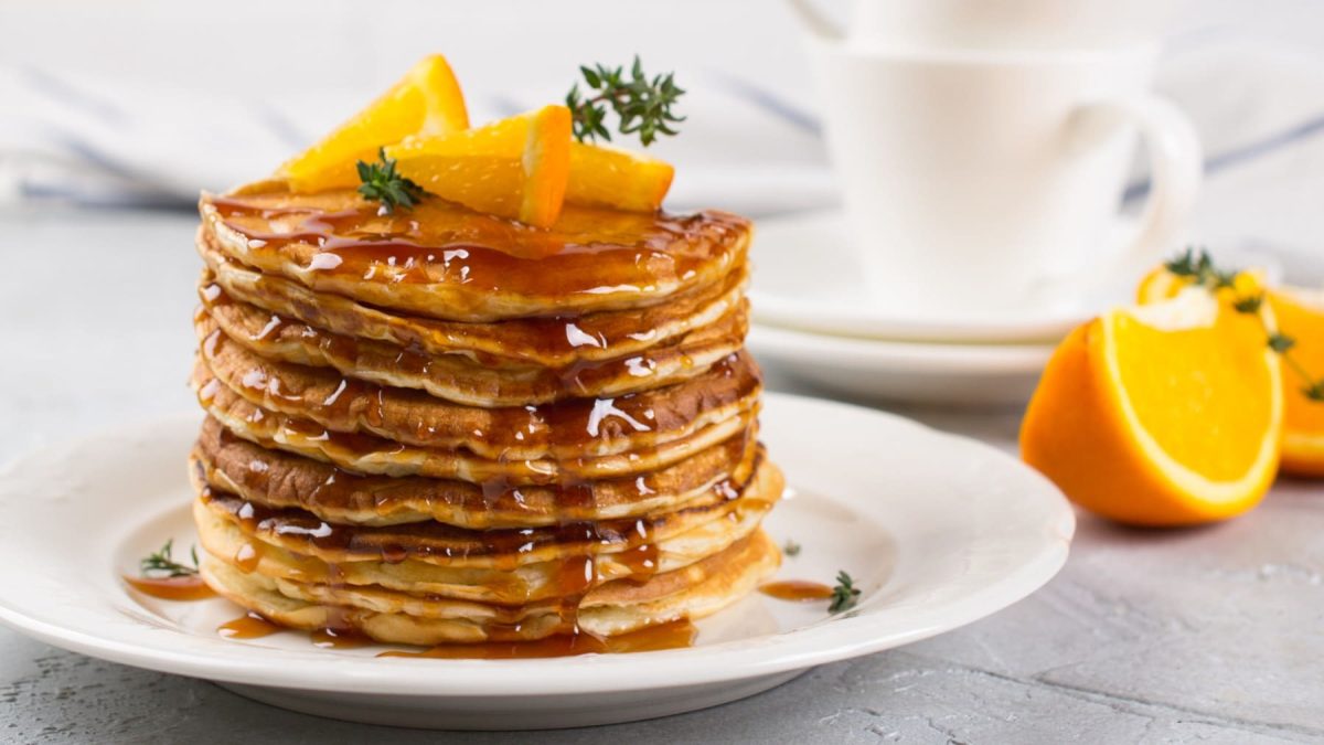 Gestapelte Orangenpancakes mit Ingwersirup und Orangenstückchen auf einem weißen Teller, im hellen Hintergrund Orangenspalten und Geschirr.