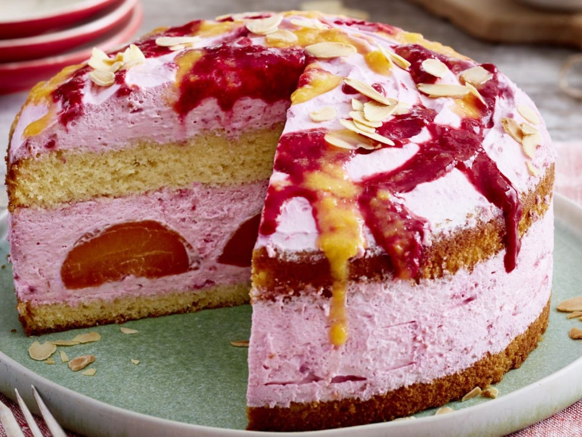 Eine angeschnittene Torte mit 4 Schichten auf einem hellgrünen Teller auf einem roten Geschirrtuch. In der Cremeschicht befinden sich halbe Pfirsiche und verziert ist die Torte mit Fruchtpürees und Mandelblättchen.