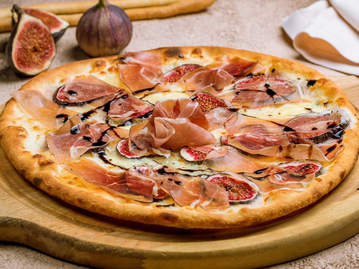 Frontal: auf einem runden Holzbrettchen liegt eine Pizza mit Feigen und Schinken. Im Hintergund liegen ausgeschnittene Feigen.
