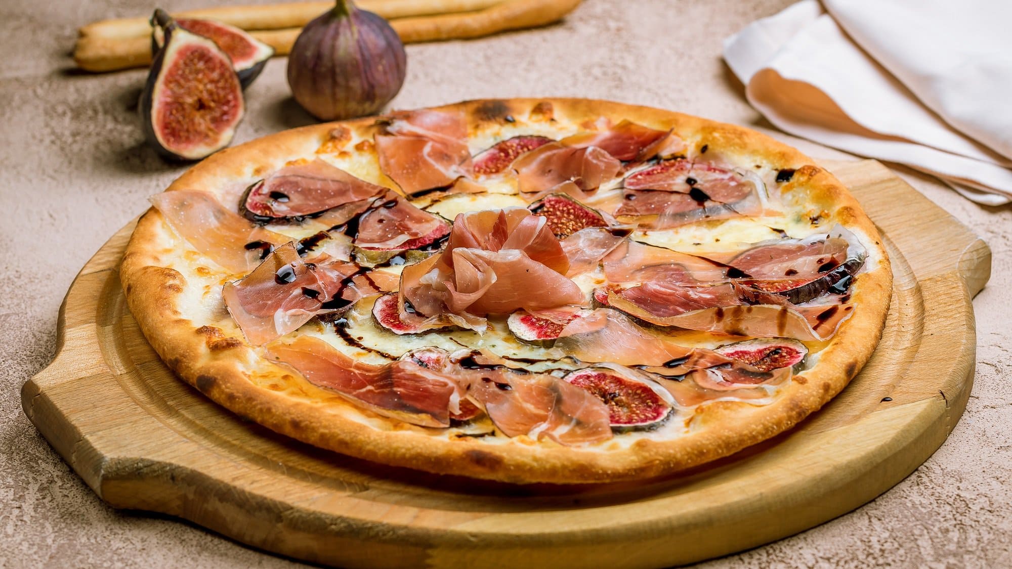 Frontal: auf einem runden Holzbrettchen liegt eine Pizza mit Feigen und Schinken. Im Hintergund liegen ausgeschnittene Feigen.