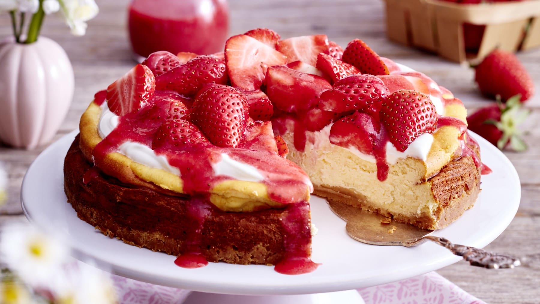 Ein Cheesecake mit Erdbeeren und ein Tortenheber auf einem weißen Teller. Links oben eine kleine Vase mit Blumen, rechts oben eine Packung Erdbeeren.