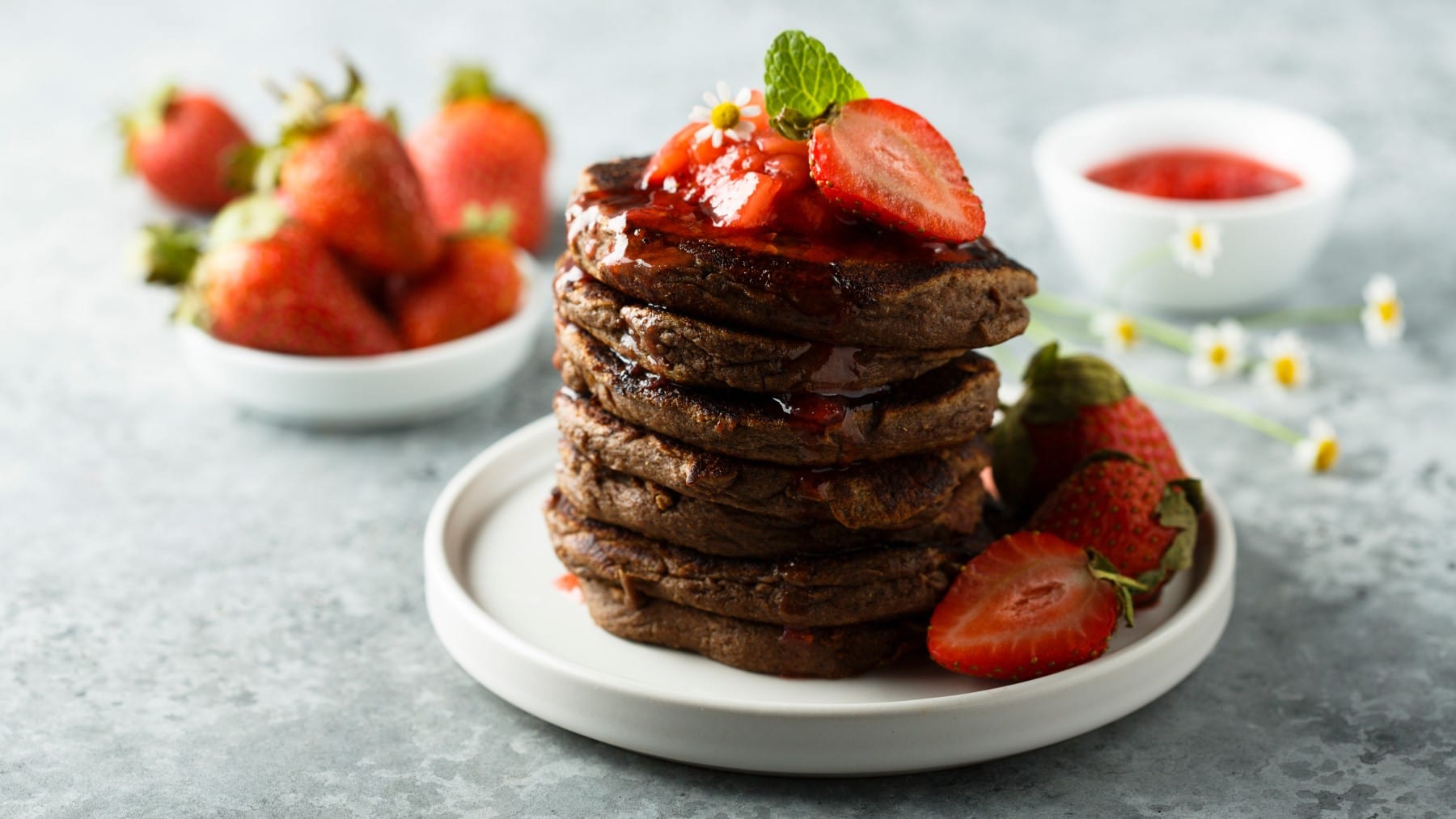 Vegane Schoko-Pancakes mit Erdbeersauce und frischen Erdbeeren auf einem weißen Teller, im Hintergrund mehr Erdbeeren, alles auf hellgrauem Untergrund.