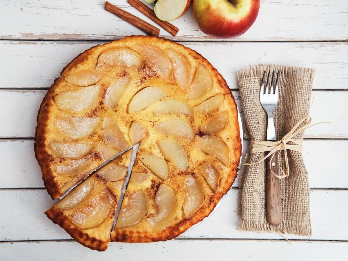 Ein angeschnittener veganer Apfelkuchen in der Draufsicht, daneben eine Gabel und frische Äpfel.
