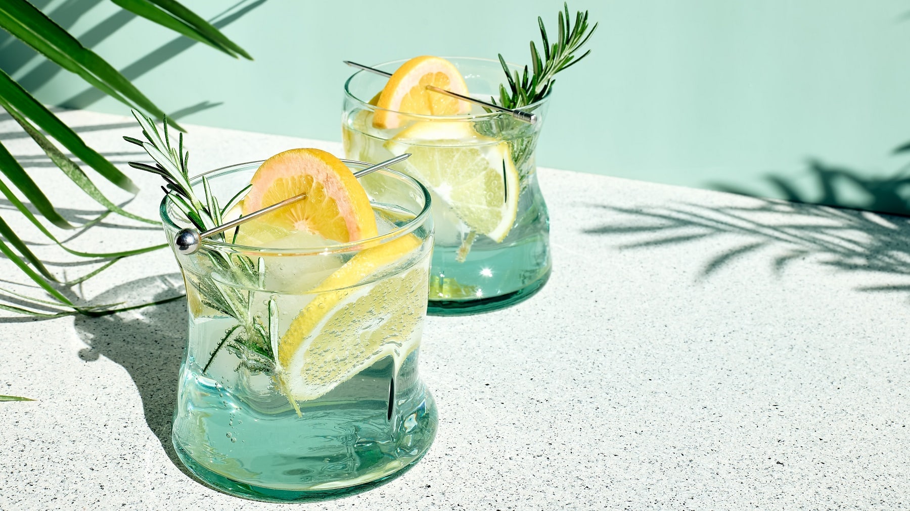 Zwei Gläser mit dem Vodka Tonic auf einem hellen Steintisch mit grünem Hintergrund und Palmen links und rechts.