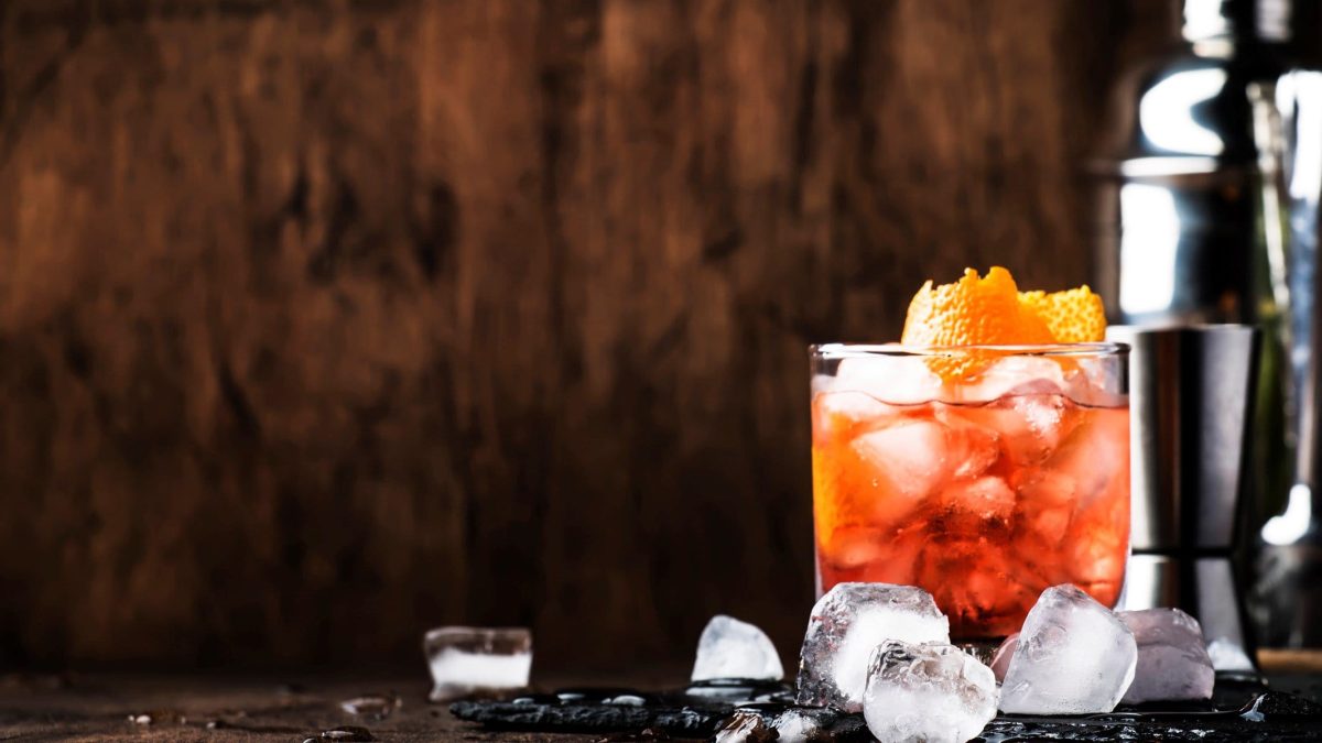 Americano Cocktail mit Orangenschale im Glas mit Eiswürfeln vor dunklem Hintergrund. Im Bild Eiswürfel und ein Cocktailshaker. Frontalansicht.