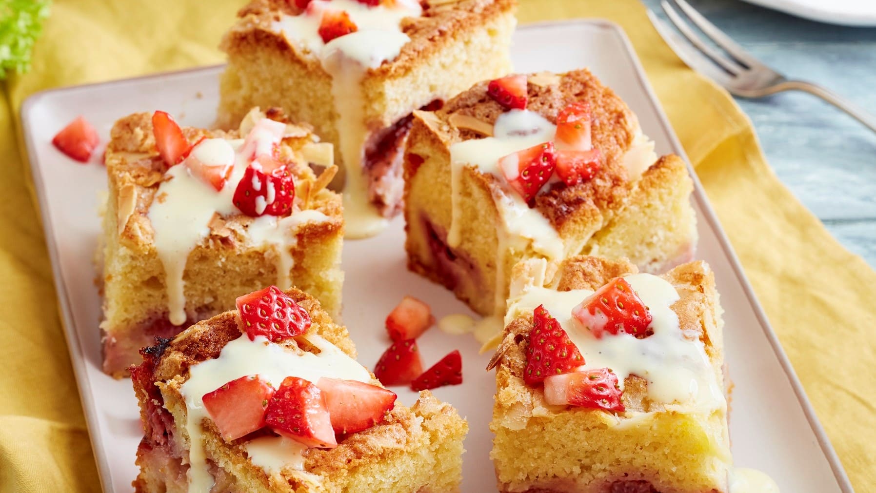 Angeschnittener Erdbeer-Pudding-Butterkuchen in Vierecken mit Guss und frischen Erdbeeren drauf auf einem beigen Teller auf Holzuntergrund.