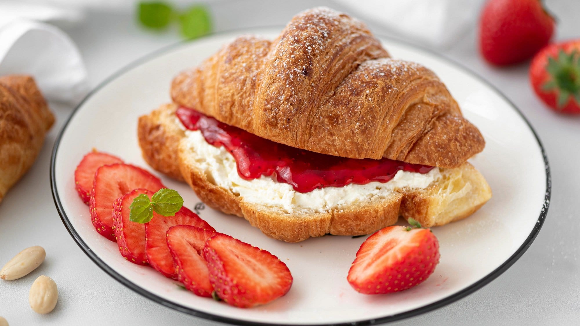 Auf einem weißen Teller liegt ein Erdbeer-Croissant mit Ricotta-Minze. Neben dem Gebäck liegen mehrere Erdbeerscheiben.