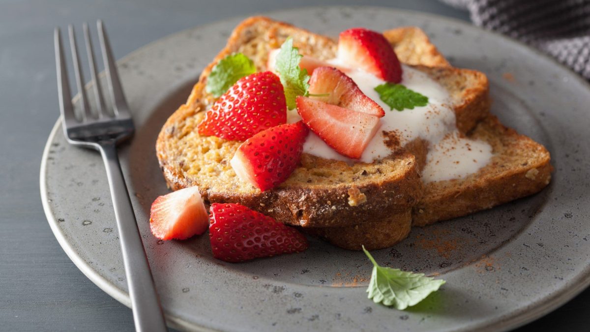 In Ahornsirup getränkter French Toast mit Erdbeeren und Vanillejoghurt auf Teller mit Gabel und Minze auf grauem Untergrund. Draufsicht.