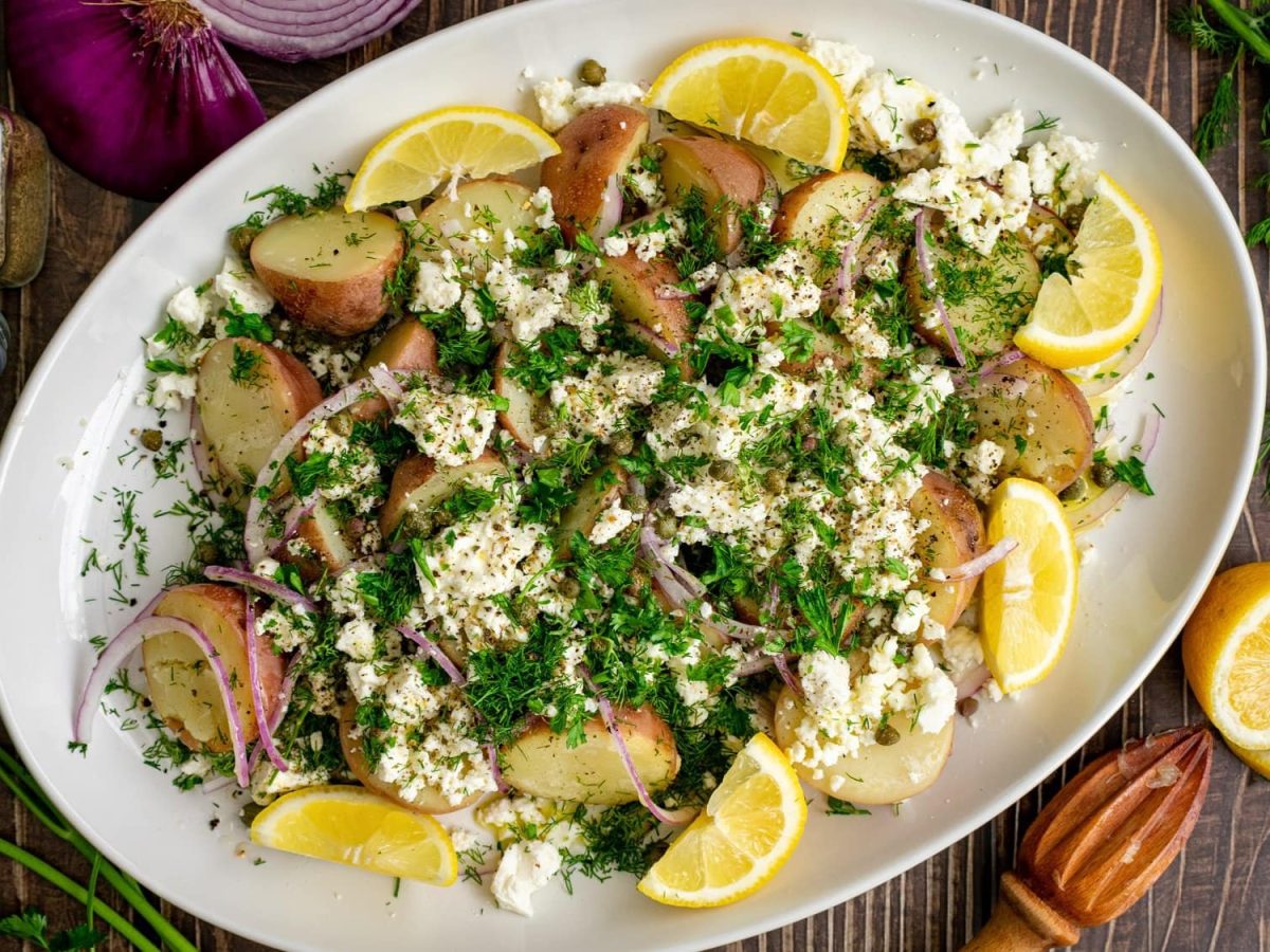 Ein Teller griechischer Kartoffelsalat mit Feta, roten Zwiebeln und Zitronenspalten sowie frischen Kräutern. Draufsicht.