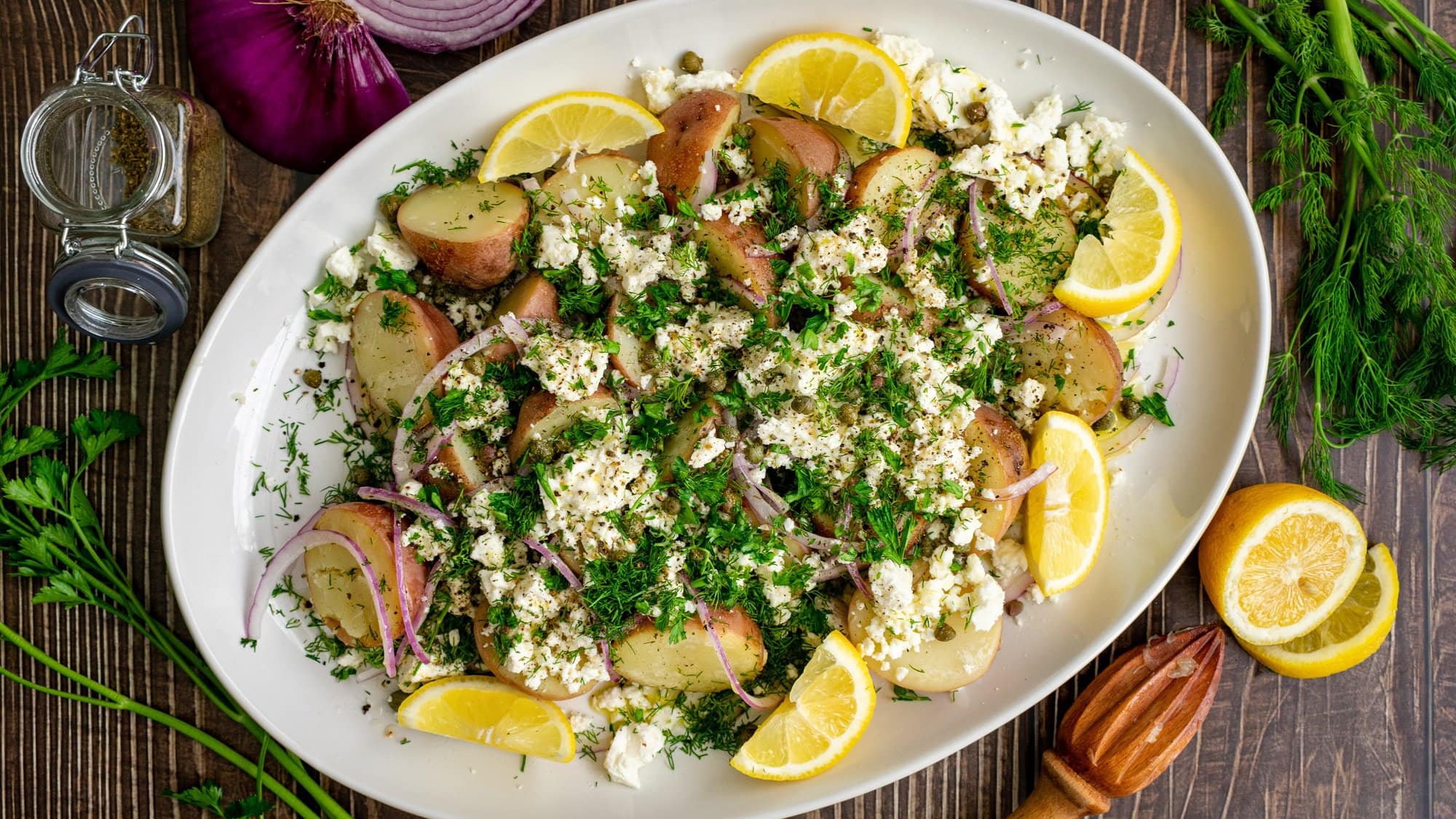 Ein Teller griechischer Kartoffelsalat mit Feta, roten Zwiebeln und Zitronenspalten sowie frischen Kräutern. Draufsicht.