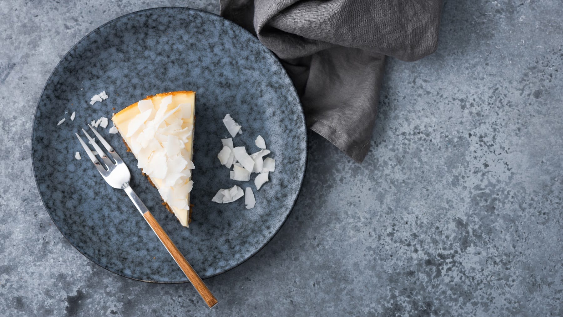 Ein blauer Tisch mit einem grauen Teller und einem Stück vom Kokosnuss-Käsekuchen mit einer Kuchengabel direkt von oben fotografiert.
