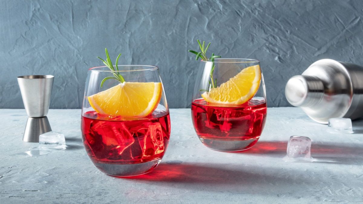 Was ist der Unterschied zwischen Wermut und Martini zwei Gläser mit rotem Wermut, Orange und Eis auf hellem Untergrund. Im Bild Barwerkzeuge. Frontalansicht.