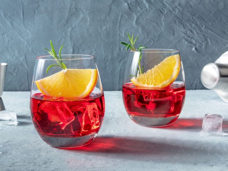 Was ist der Unterschied zwischen Wermut und Martini zwei Gläser mit rotem Wermut, Orange und Eis auf hellem Untergrund. Im Bild Barwerkzeuge. Frontalansicht.