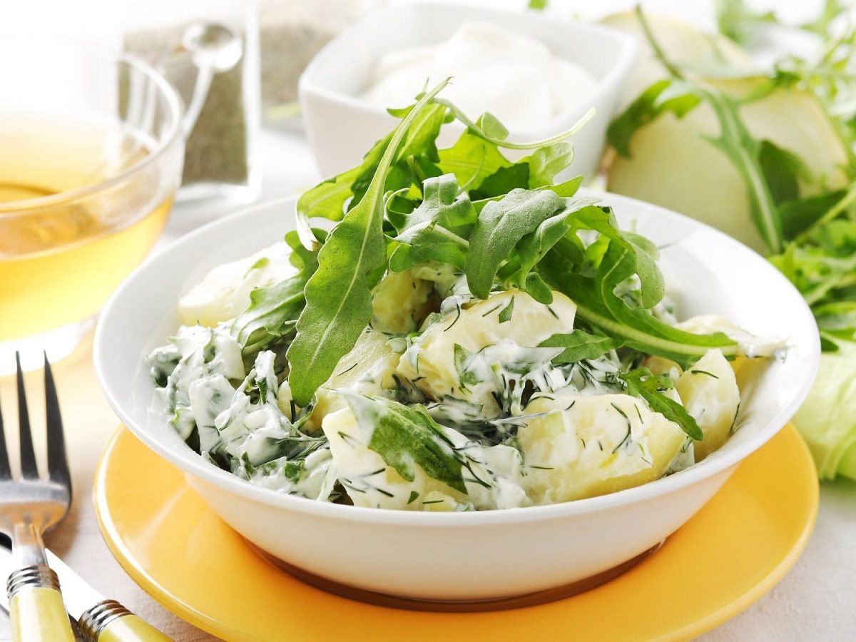 Eine Schale Rucola-Kartoffelsalat mit griechischem Joghurt und Dill, daneben eine Gabel und Zutaten.