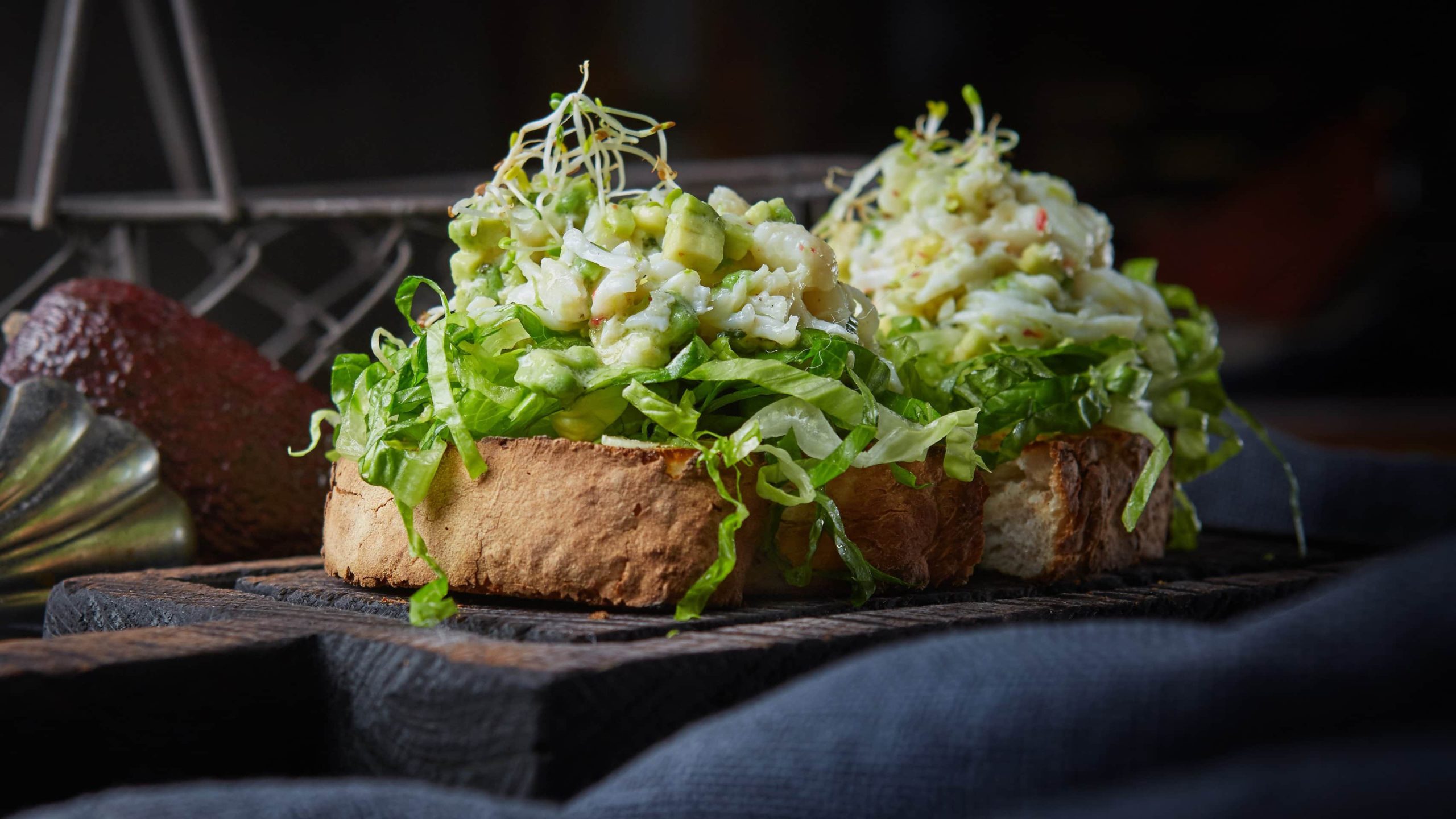 Ein dunkler Hinter-und Untergrund. In der Mitte das Sandwich mit dem Avocado-Garnelen-Eiersalat seitlich fotografiert.
