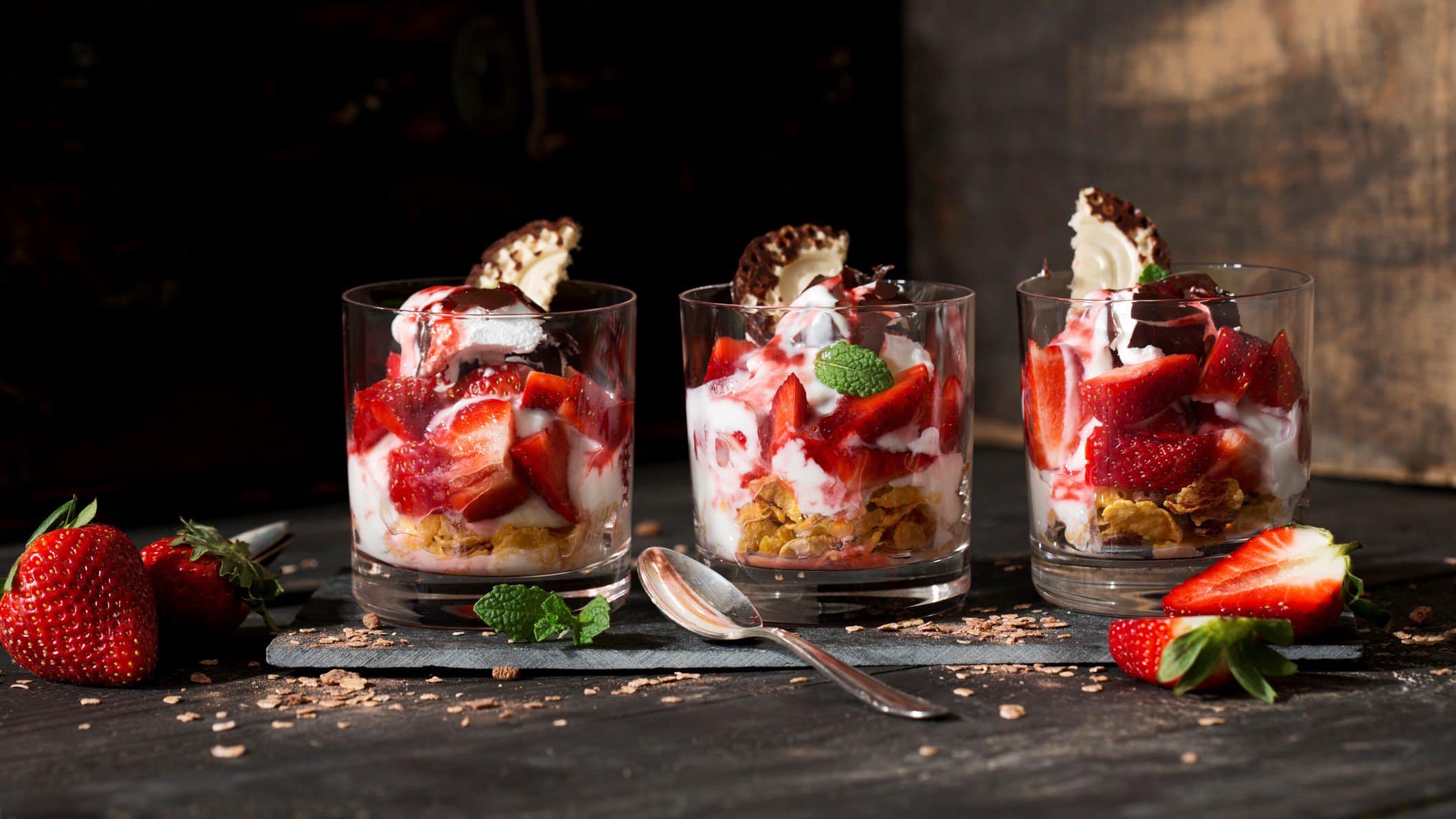Schokokuss-Erdbeer-Dessert in drei durchsichtigen Gläsern vor rustikalem Ambiente.