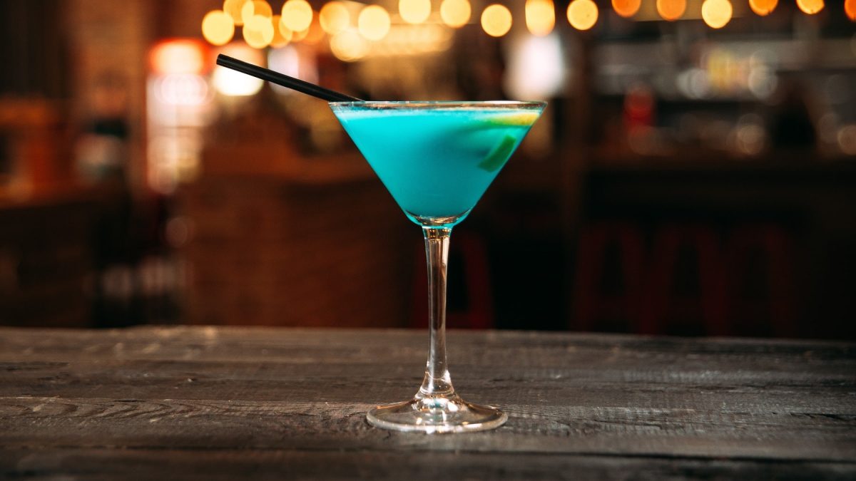 Ein Glas mit dem Swimming Pool Martini auf einer Holztheke und verschwommenen Lichtern im Hintergrund.