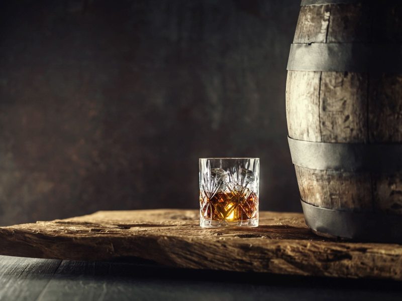 Zum Tag des Bourbon Was ist Bourbon? Ein Glas mit Bourbon Whiskey und Holzfass auf Holz vor grauem Hintergrund. Frontalansicht.
