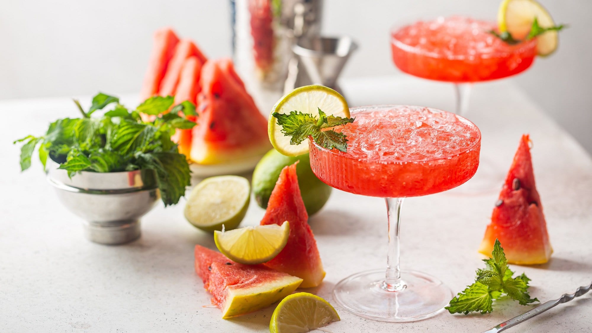Zwei Gläser Wassermelonen-Daiquiri, darum frische Wassermelone und MInze sowie Cocktail-Zubehör.
