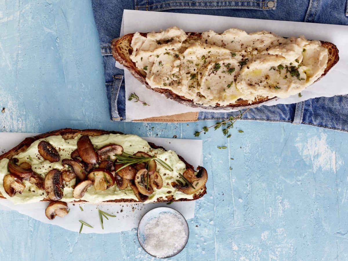 Zweierlei getoastetes Hirnfutter: Je eine Scheibe Brot mit Bohnenmus und Avocado und Pilzen auf weißen Unterlagen, darunter blaue Fläche.