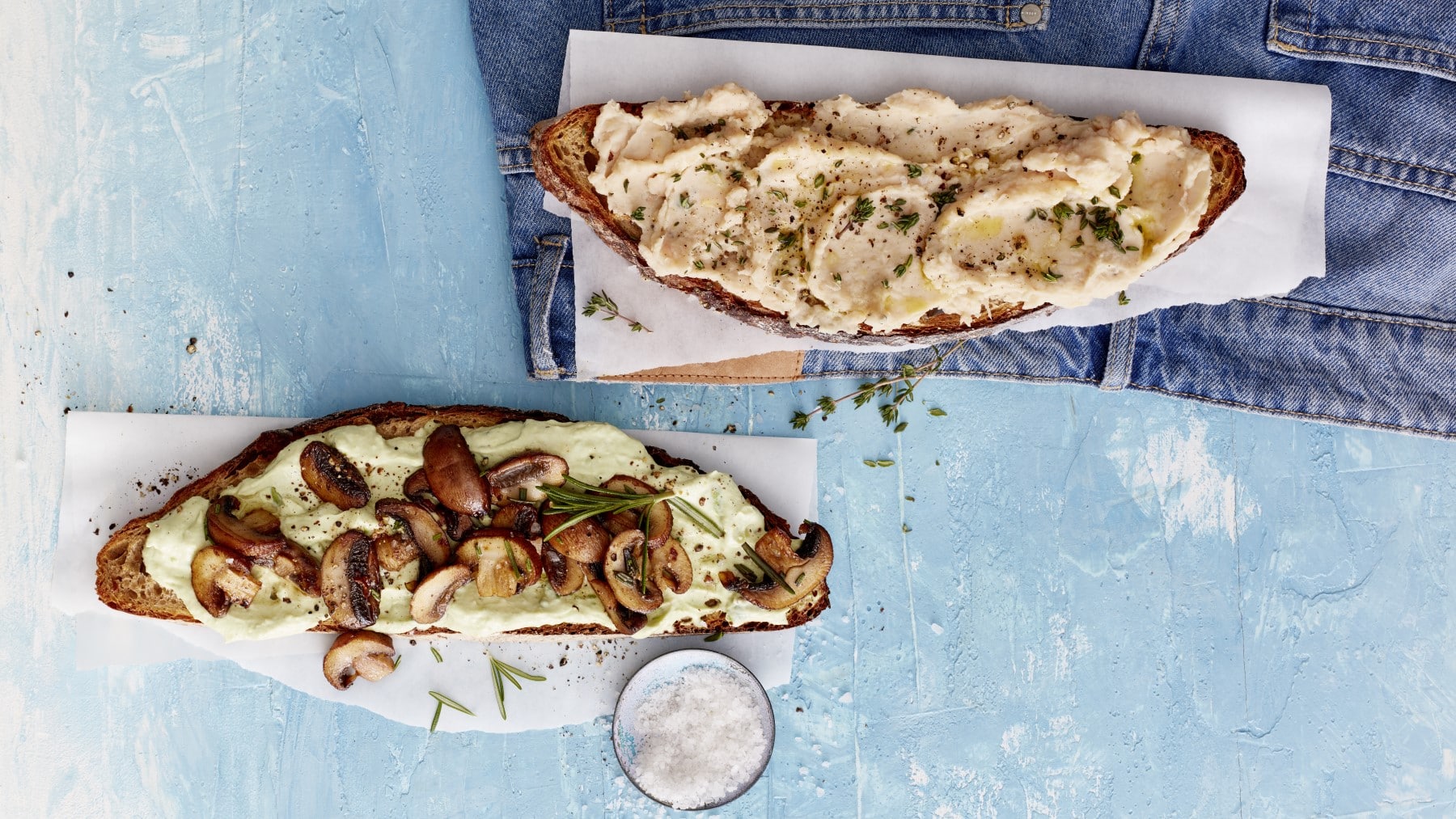 Zweierlei getoastetes Hirnfutter: Je eine Scheibe Brot mit Bohnenmus und Avocado und Pilzen auf weißen Unterlagen, darunter blaue Fläche.